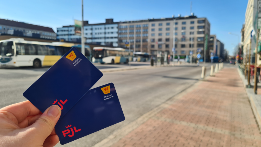 kaksi Porin joukkoliikenteen matkakorttia sormissa ja taustalla kauppatorin laiturialue jossa busseja